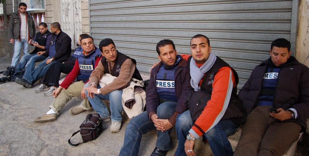 A row of men sitting on the ground against a building, several wearing vests that read PRESS