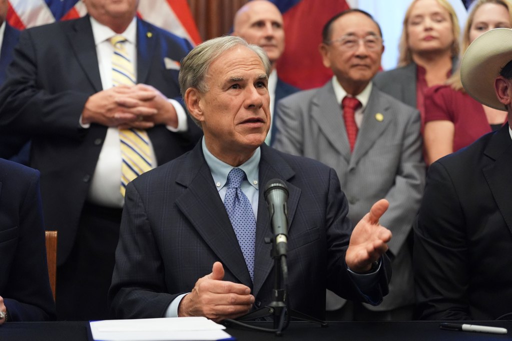 Greg Abbott speaks at a desk as legislatures stand all around him