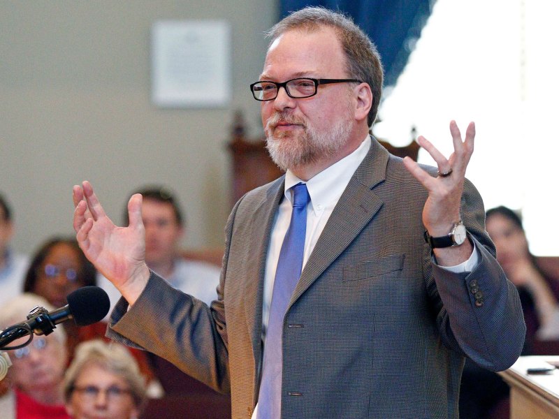 Greg Isles, in a suit, speaks at a gathering, hands wide in the air