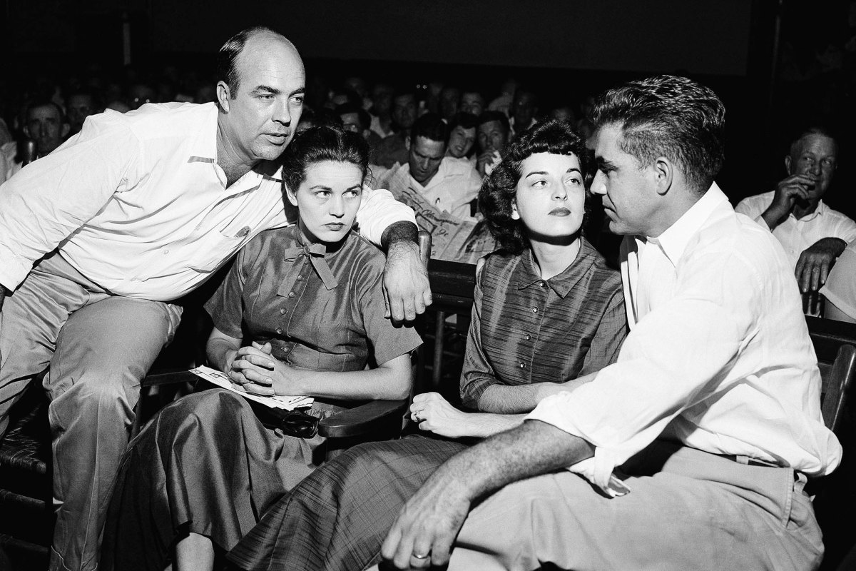 a black and white photo shows two white men sitting on either side of two women with their arms around the women in a courtroom. The clothes and hair styles are 1950s style