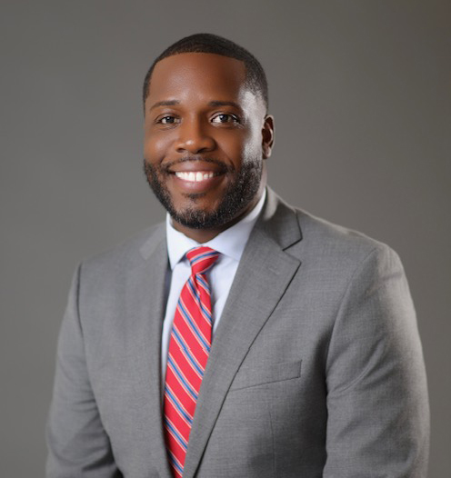 An official portrait of a bearded man in a light grey suit and wearing a red and blue stripped tie