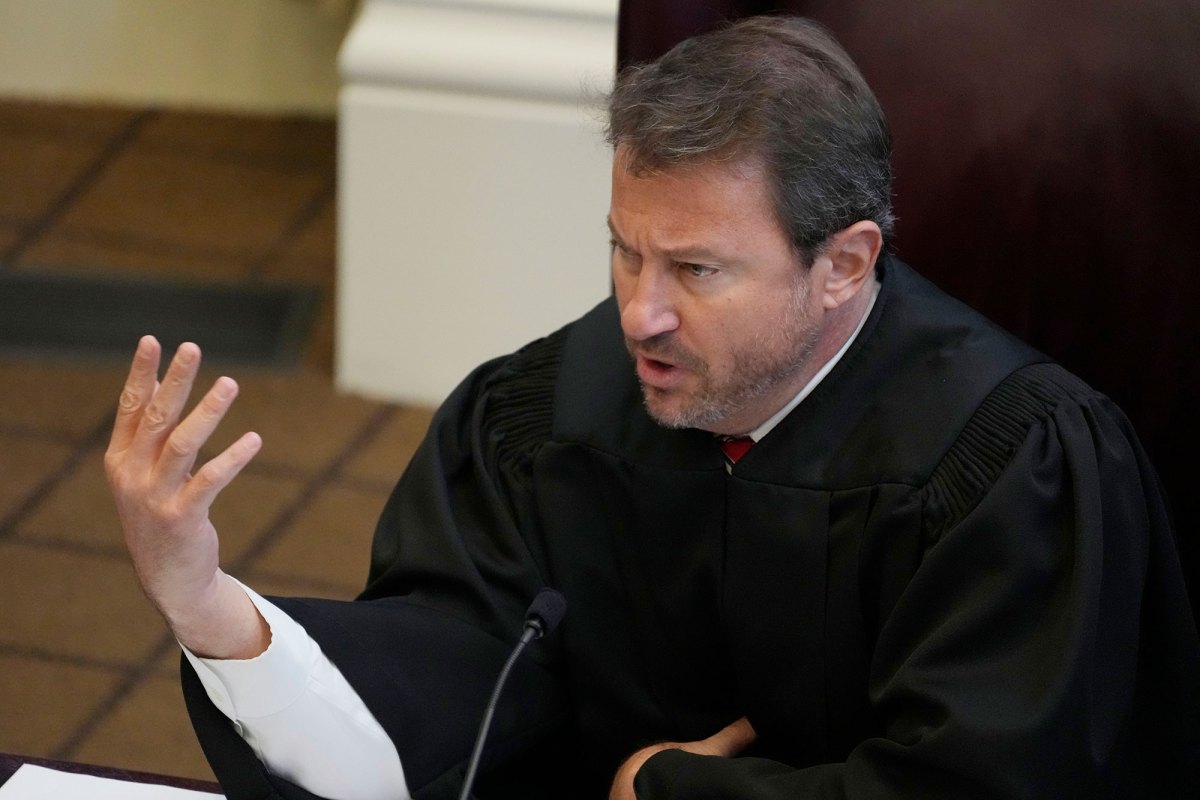 A judge, in black robes, gestures with one hand while speaking from the bench