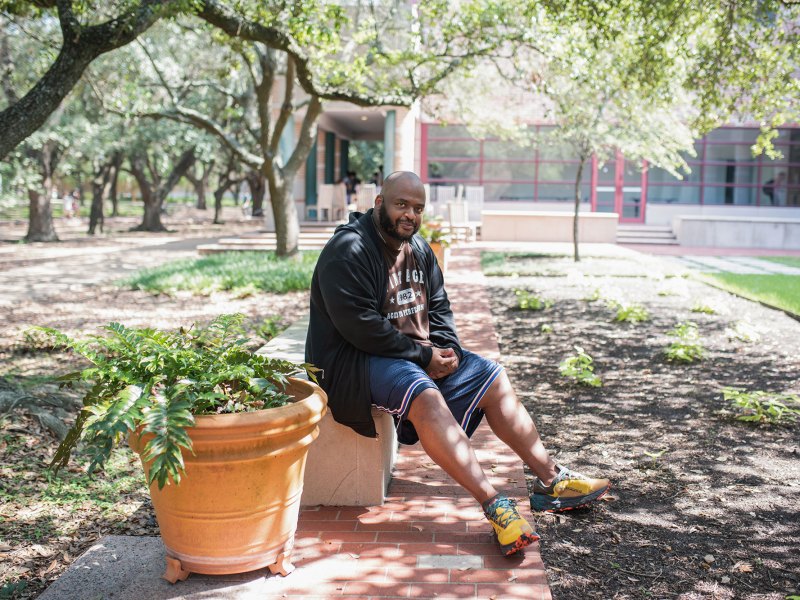 A bearded man in a hoodie and shorts sits on a bench outside in the shade of a large tree