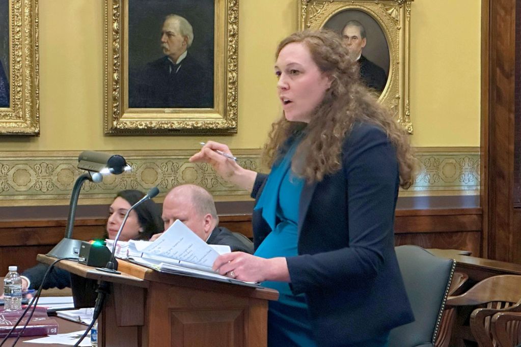 A woman with long hair stands at a podium in court, talking and gesturing with her hands
