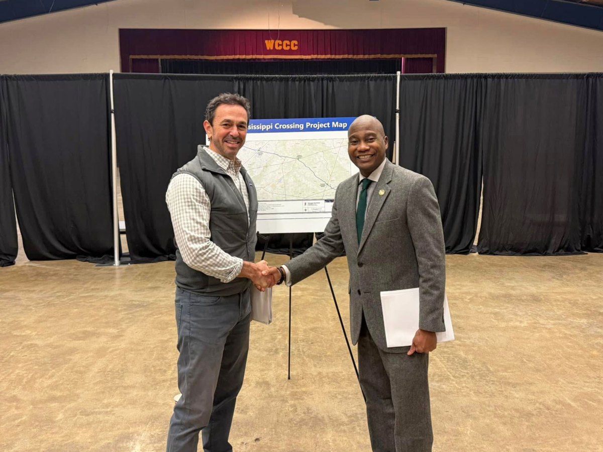 Two men shape hands in a school auditorium. A sign for Mississippi Crossing Project Map stands behind them
