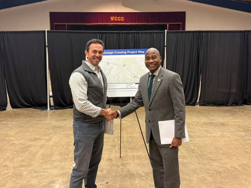 Two men shape hands in a school auditorium. A sign for Mississippi Crossing Project Map stands behind them