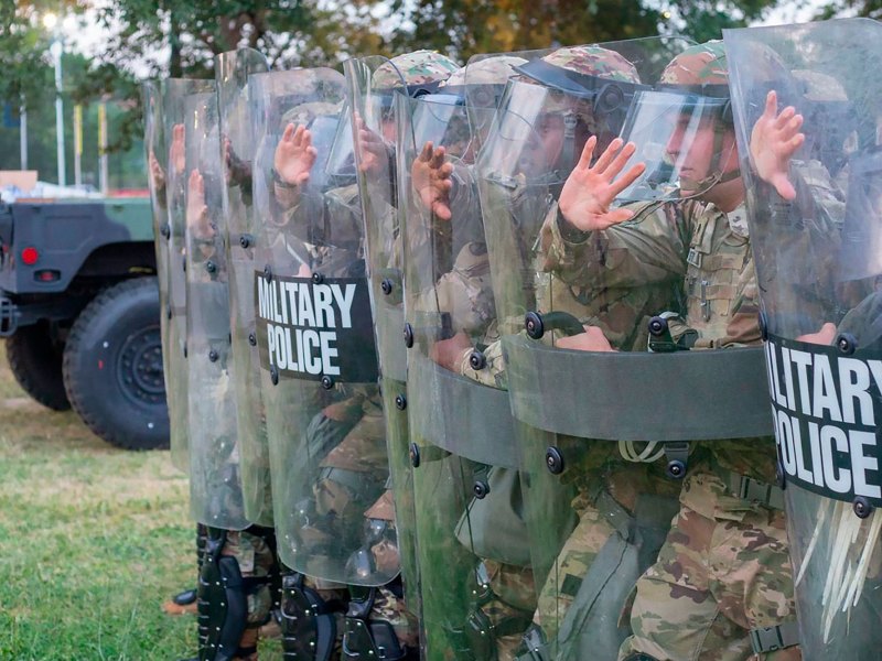 A row of men in camouflage uniforms hold up shields that read 'Military Police'