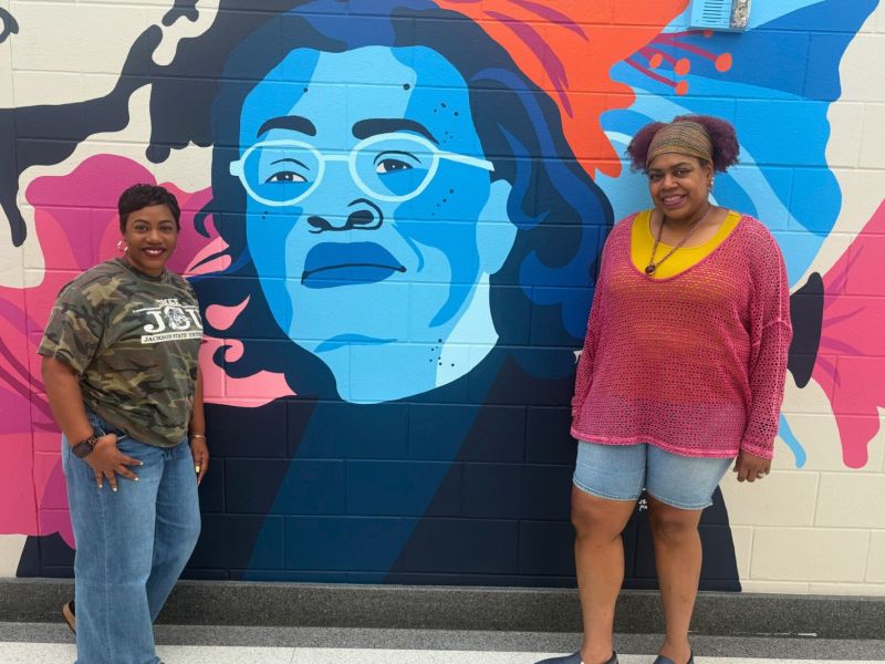 Two women stand beside a mural of a woman painted in blue