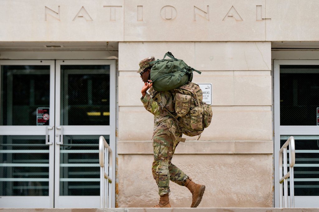 A person in military camo carries backpacks past a building labeled National
