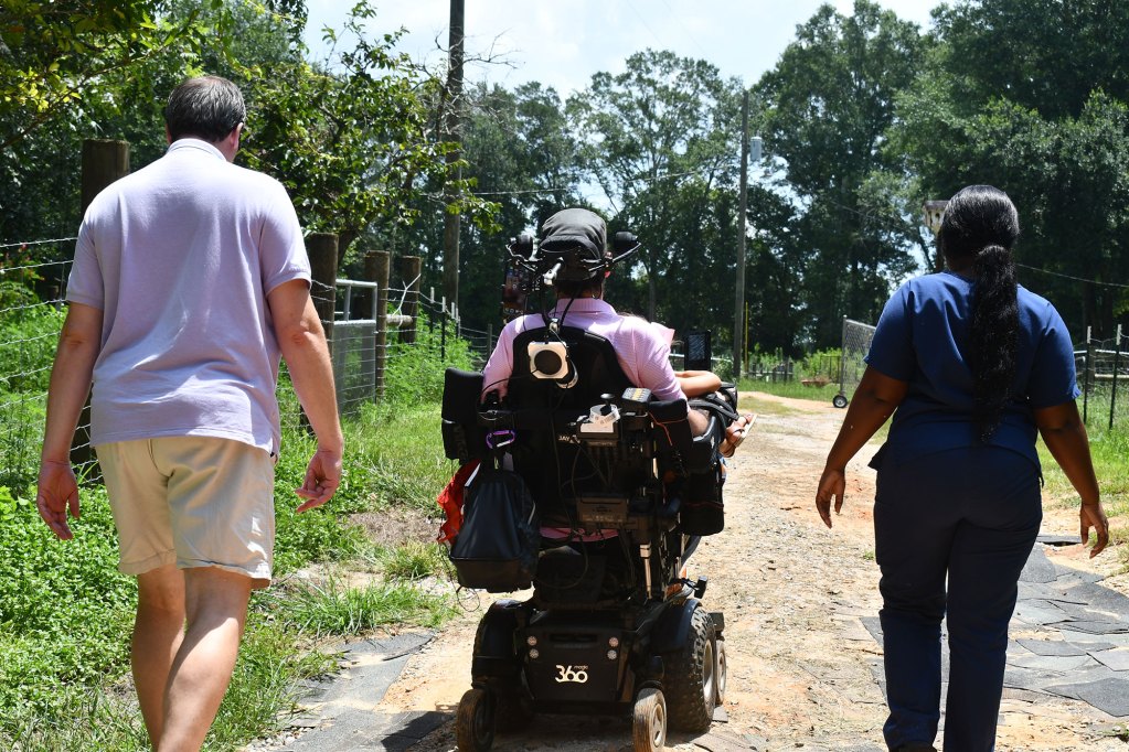 A man in a motorized wheelchair rides between two people on a rural path lined with trees and fencing.