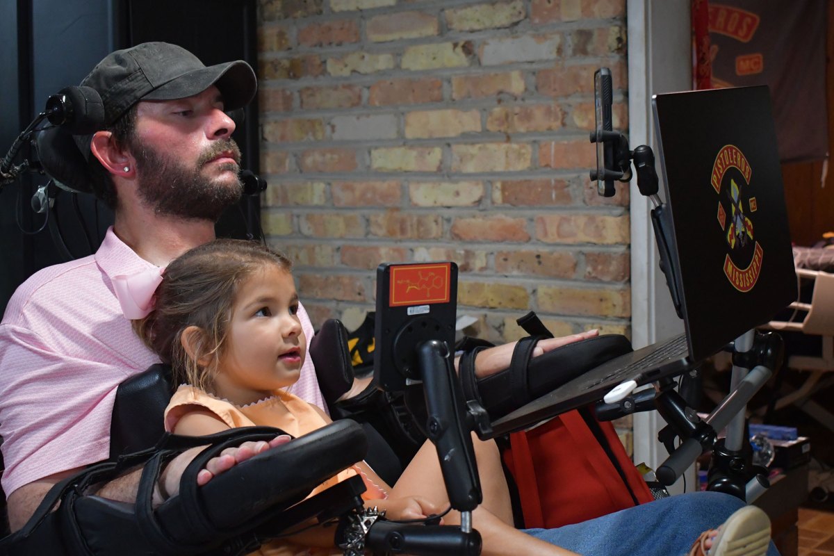 A man in a wheelchair sits with a young girl on his lap, both looking at a laptop screen.