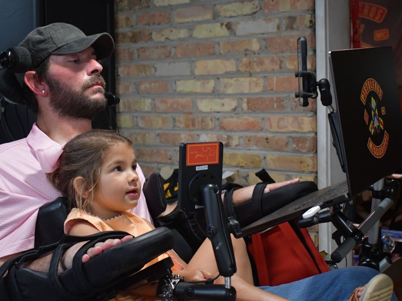 A man in a wheelchair sits with a young girl on his lap, both looking at a laptop screen.