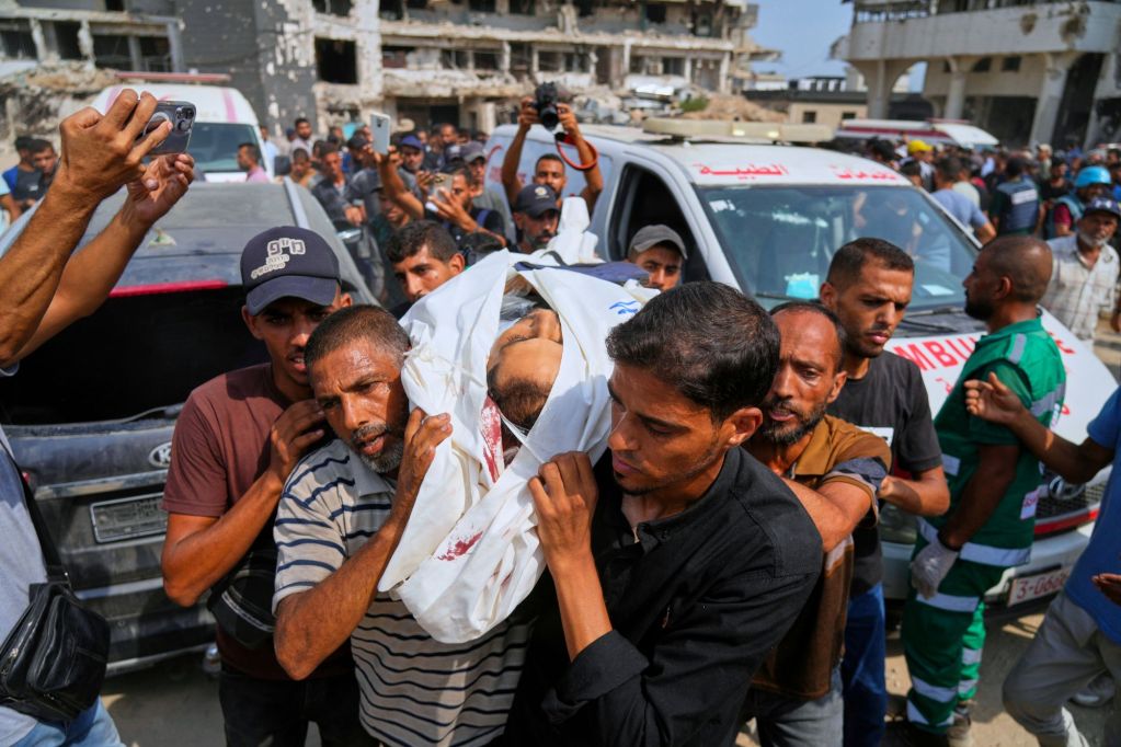 A crowd carries a body wrapped in a white sheet down a war torn road