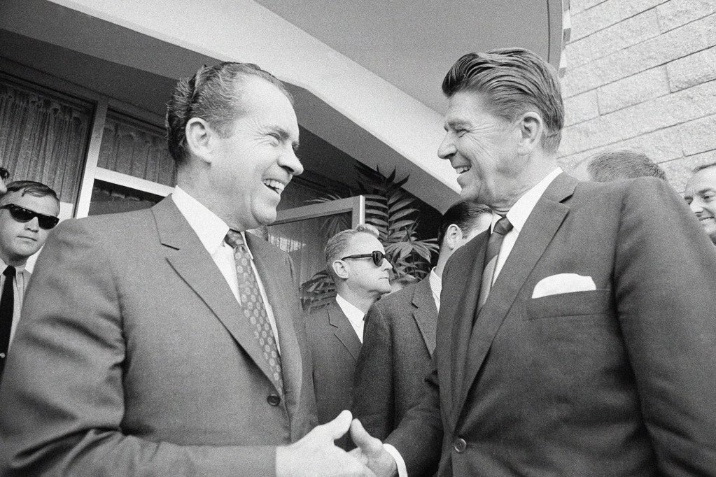 Richard Nixon and Ronald Reagan shake hands outside of a building in an old black and white photo