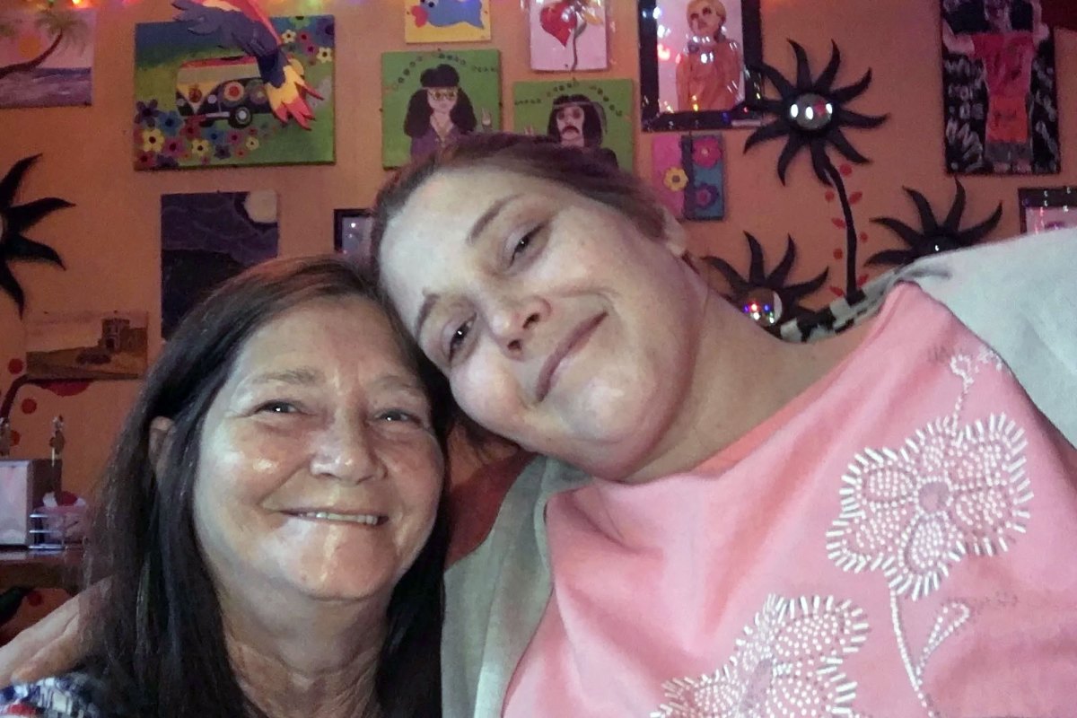 Two women lean their heads together in front of a wall covering in artwork.
