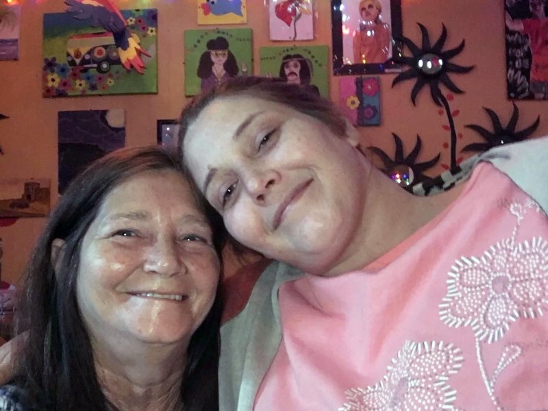 Two women lean their heads together in front of a wall covering in artwork.