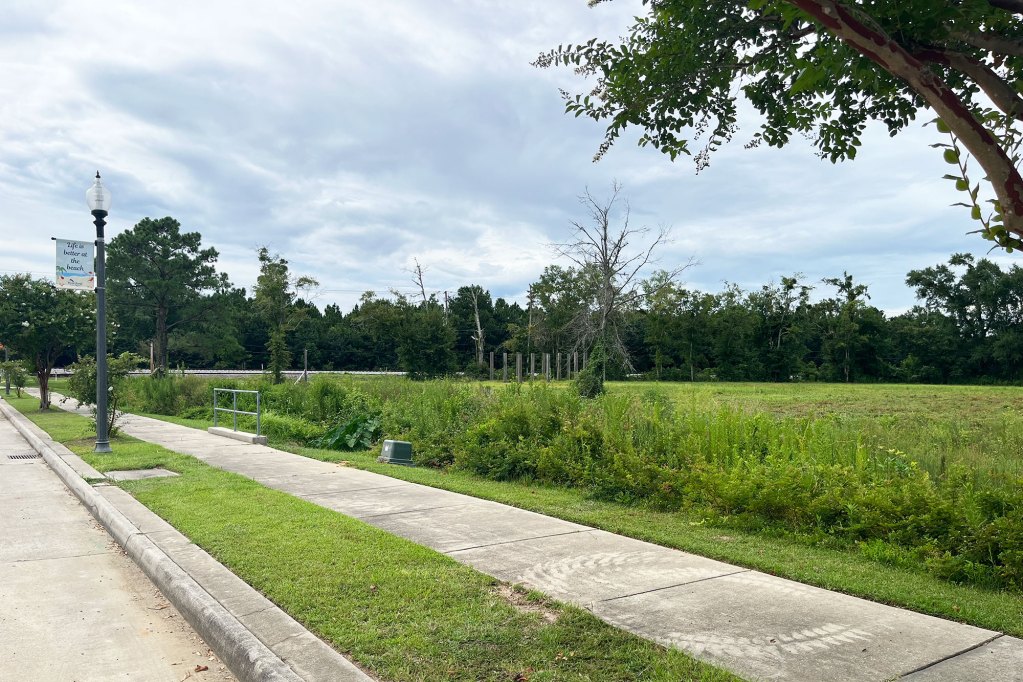 A city sidewalk goes past an empty green lot
