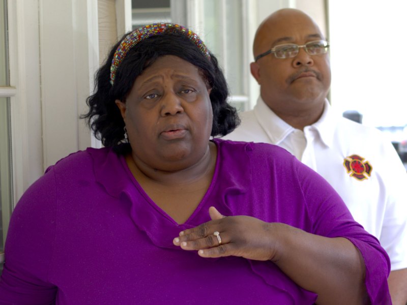 A woman in a purple top speaks outside on a covered porch as an official stands behind her
