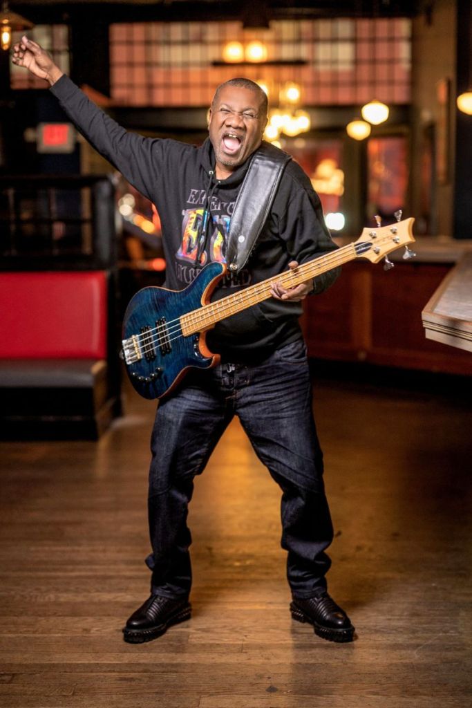A man dressed in black yells while swinging and arm wide to play the guitar strapped around his neck