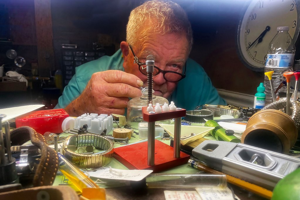 A man hunches over a work desk and looks closely at small mechanical parts