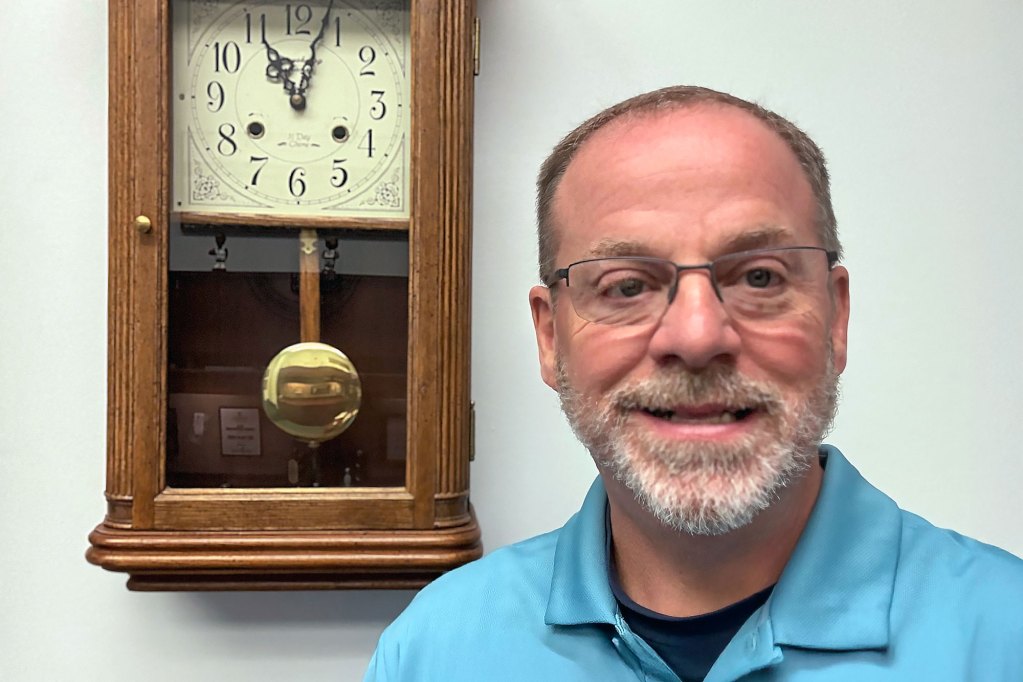 A man in a blue shirt and glasses stands in front of a wall where a clock hangs
