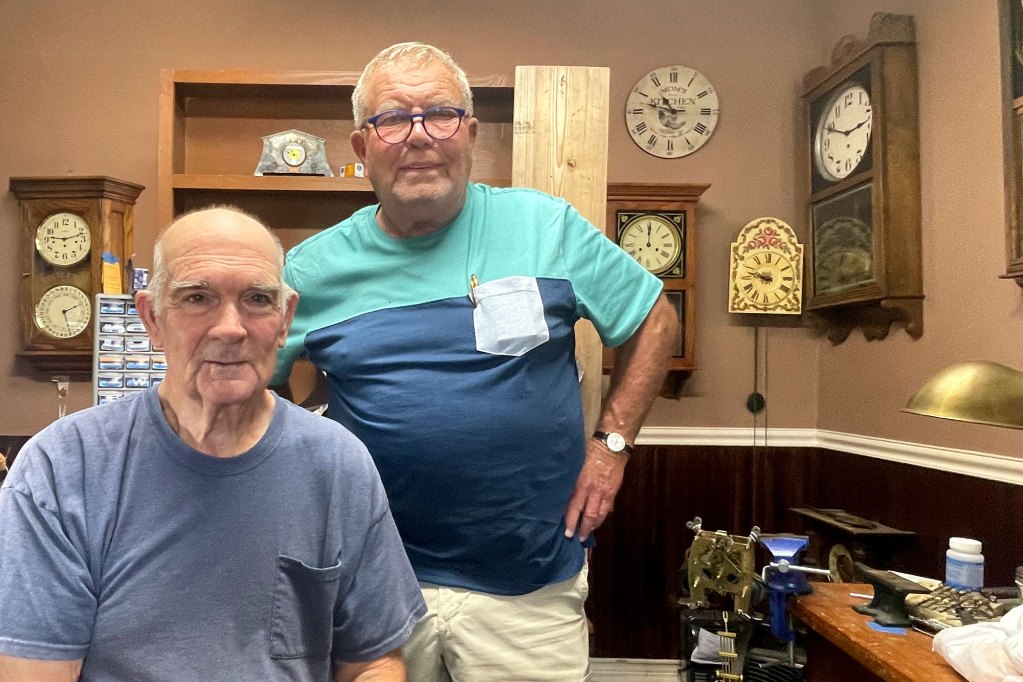 Two men in blue shirts pose inside a room with many clocks on the wall behind them