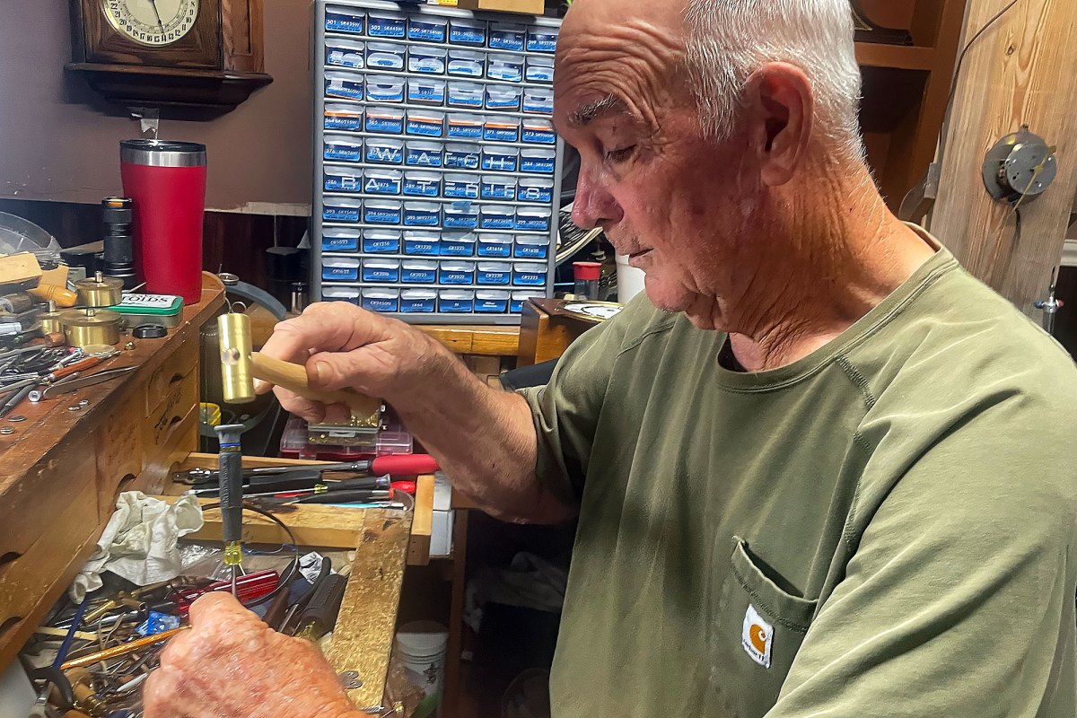 A man in a green tshirt sits at a work station and works on something with a small gold hammer