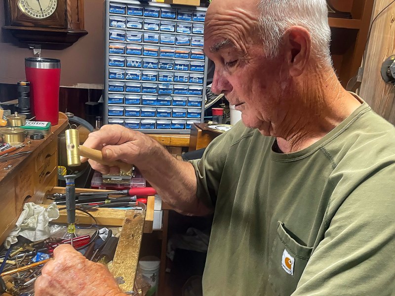 A man in a green tshirt sits at a work station and works on something with a small gold hammer