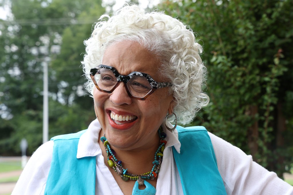 A white haired woman woman in a white top and blue vest with matching jewelry smiles outside