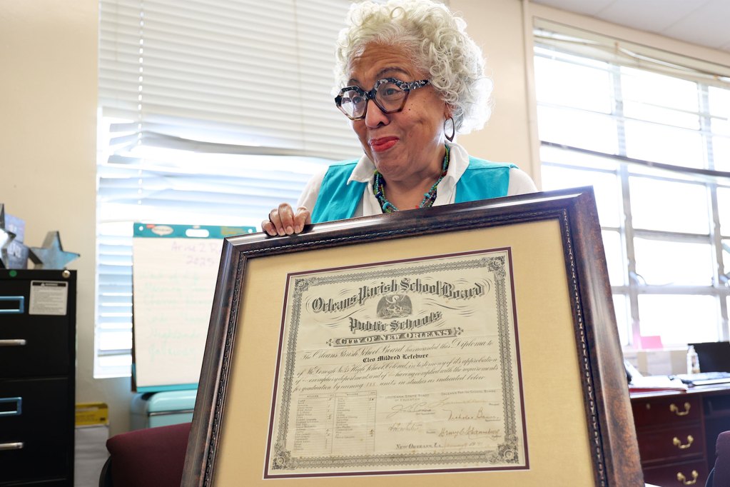A white haired woman woman in a white top and blue vest holds an old framed diploma in her office