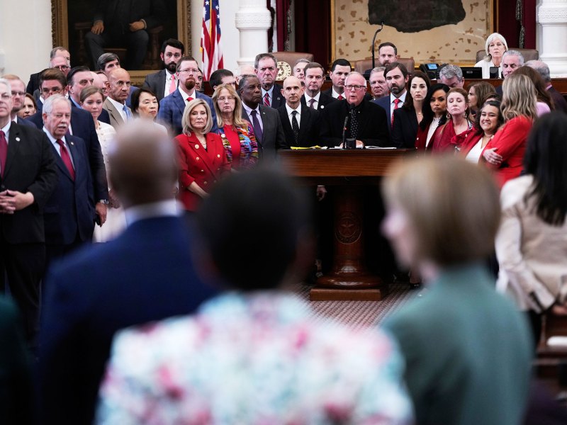 A crowd of legislatures stand together on one side of a room
