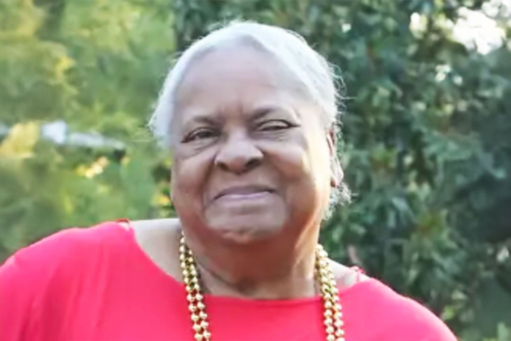 A woman in a red top and gold beads around her neck smiles outside for a photo