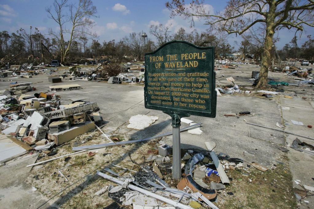 a photo shows devastation, downed trees, and slabs of concrete covered in debris around a standing green sign that reads: "FRom the People of Waveland - In appreciation and gratitude to all who gave their time, energy, and money to help us recover from Hurricane Camille. On August 17, 1969 our city was devastated but those who cared came to her rescue."
