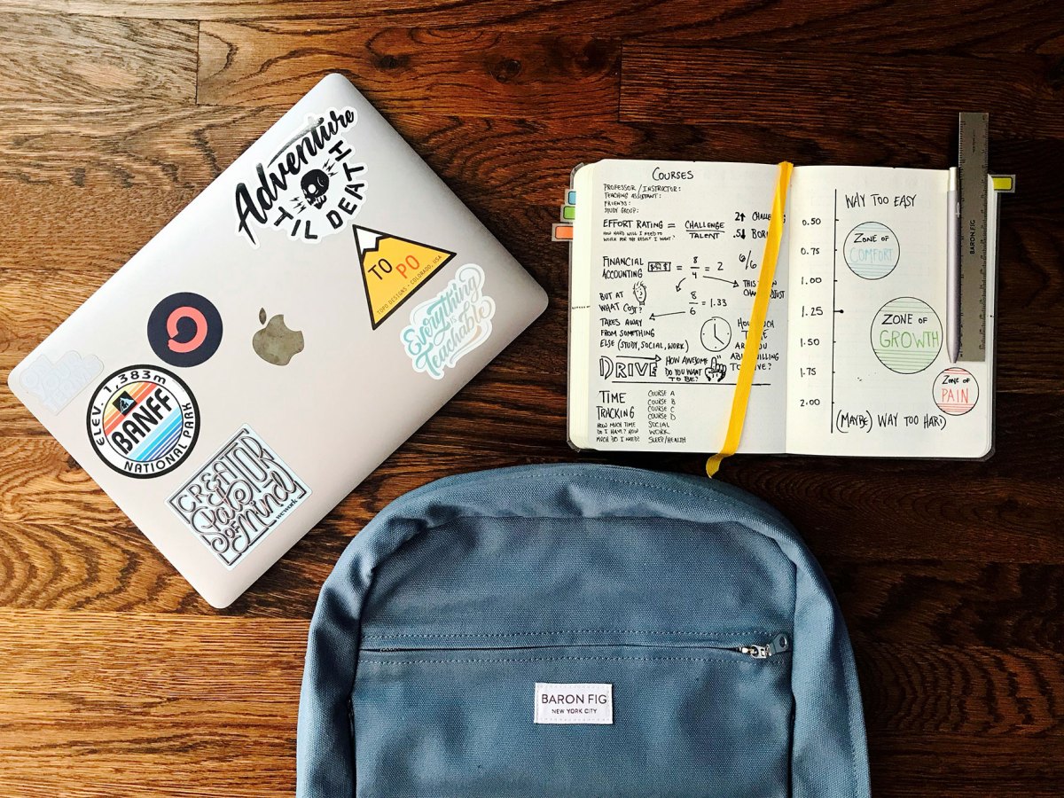 A flatlay of a blue backpack, a mac laptop covered in stickers, and an open notebook