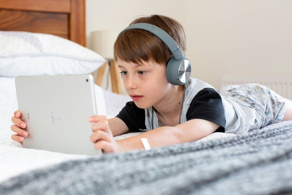 A child lays across a bed with tablet in hands and headphones on