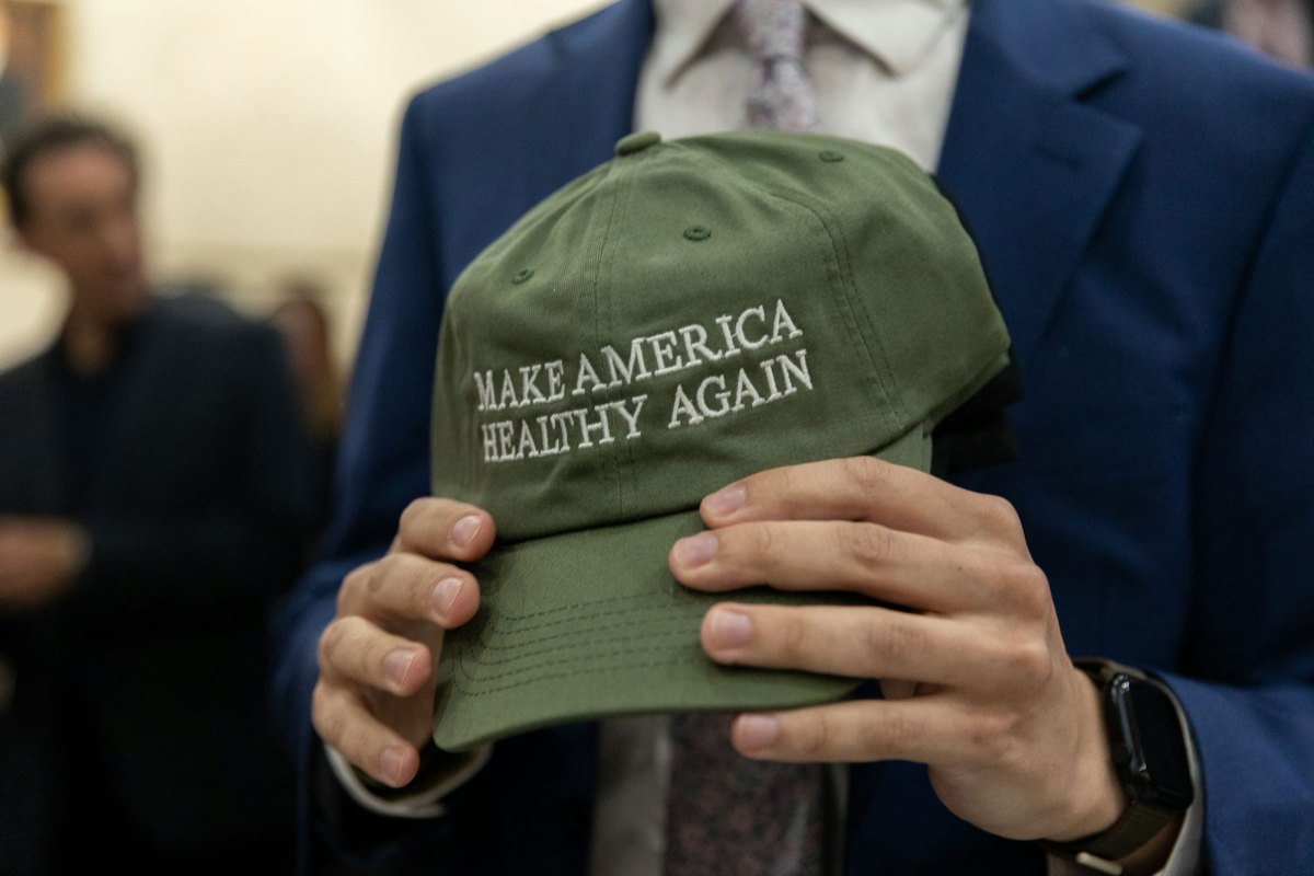 Closeup of a pair of hands holding up an olive green ball cap that reads 'MAKE AMERICA HEALTHY AGAIN'