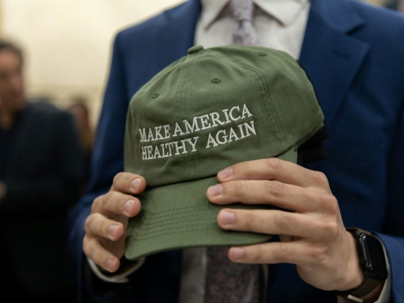 Closeup of a pair of hands holding up an olive green ball cap that reads 'MAKE AMERICA HEALTHY AGAIN'