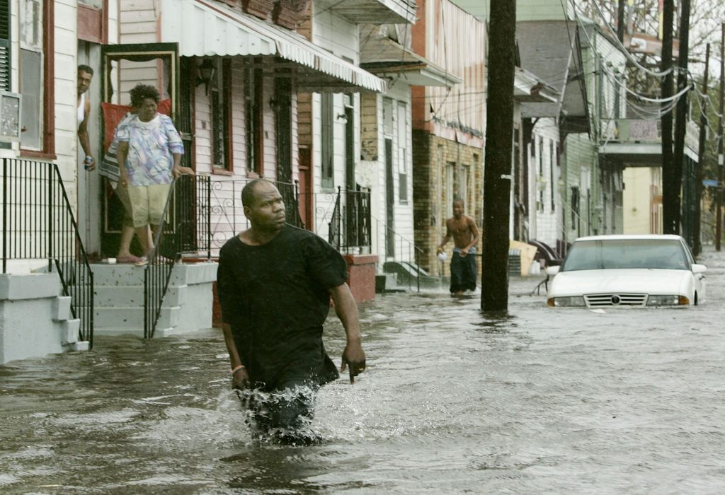 A man with a distressed look walks in thigh-deep water while people watch from a doorway several steps above street level.