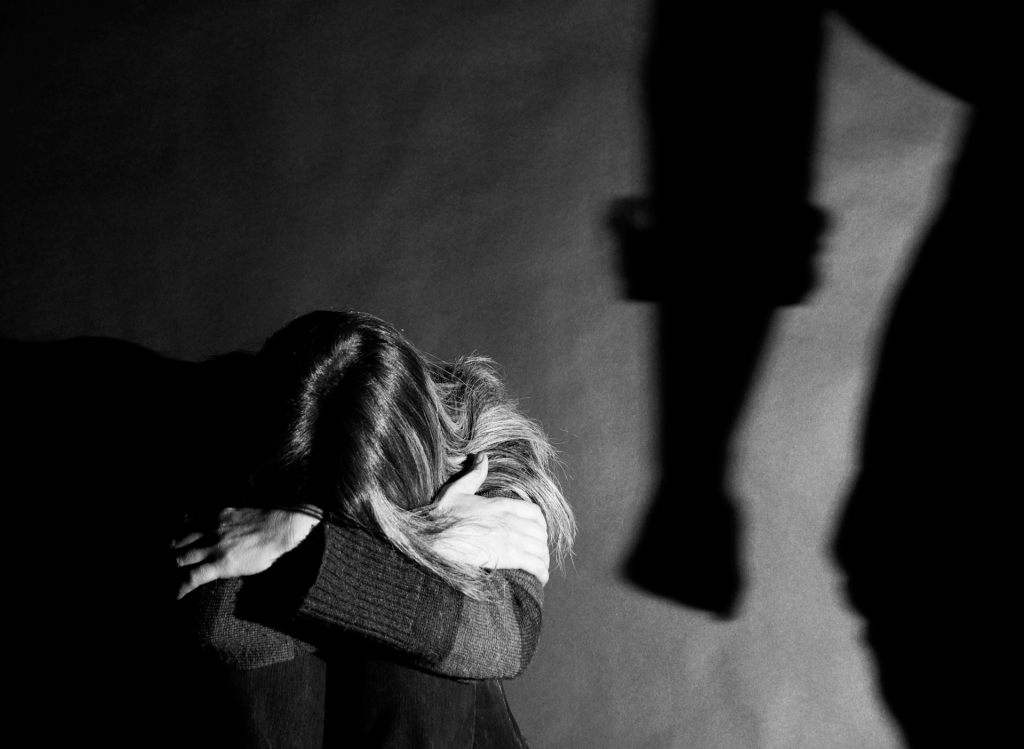 A woman on the floor, face hidden in her knees, with the looming shadow of a man’s fist beside her