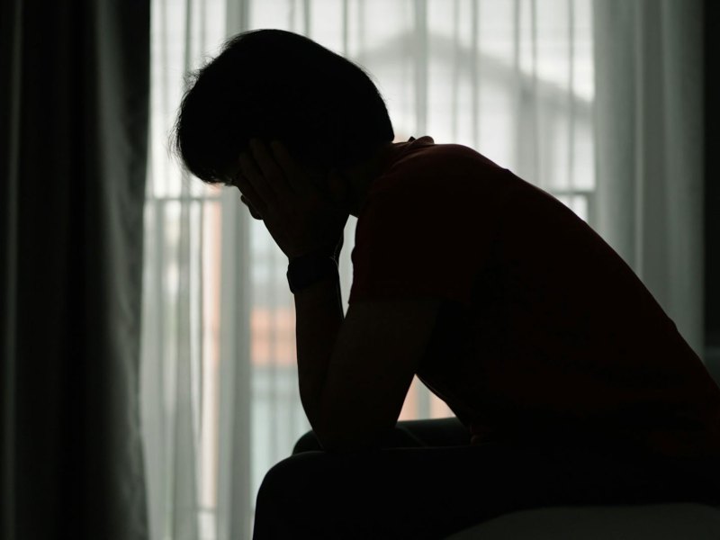 The silhouette of a woman sitting on the edge of her bed in front of a window, head in hands