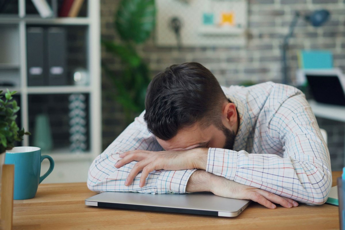 A man sitting at a desk with his head in his hands, leaning on a closed laptop
