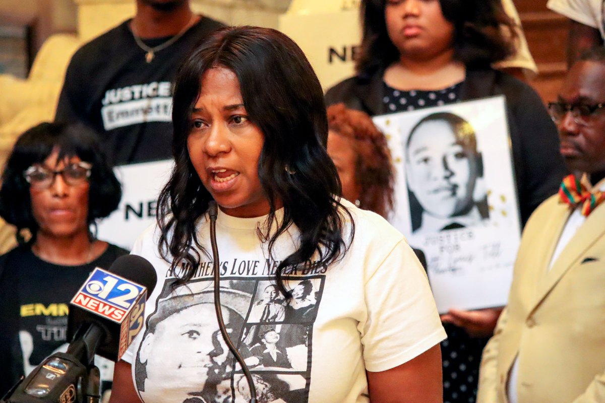 A woman wearing a screenprinted white T-shirt with pictures of Emmett Till speaks into a microphone in front of rows of people.
