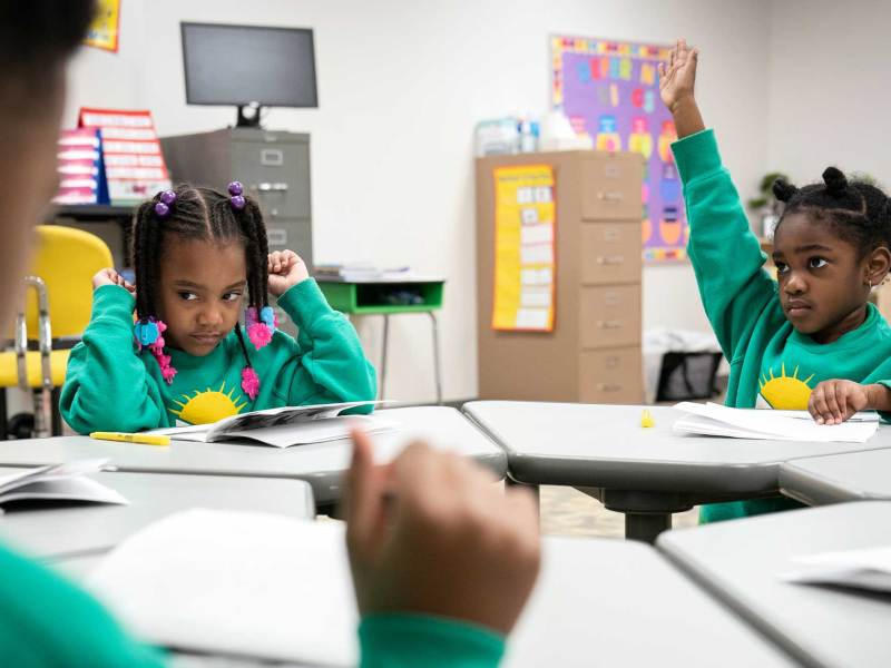 Young students in matching green sweaters sit around their tables, one raising her hand for the teacher