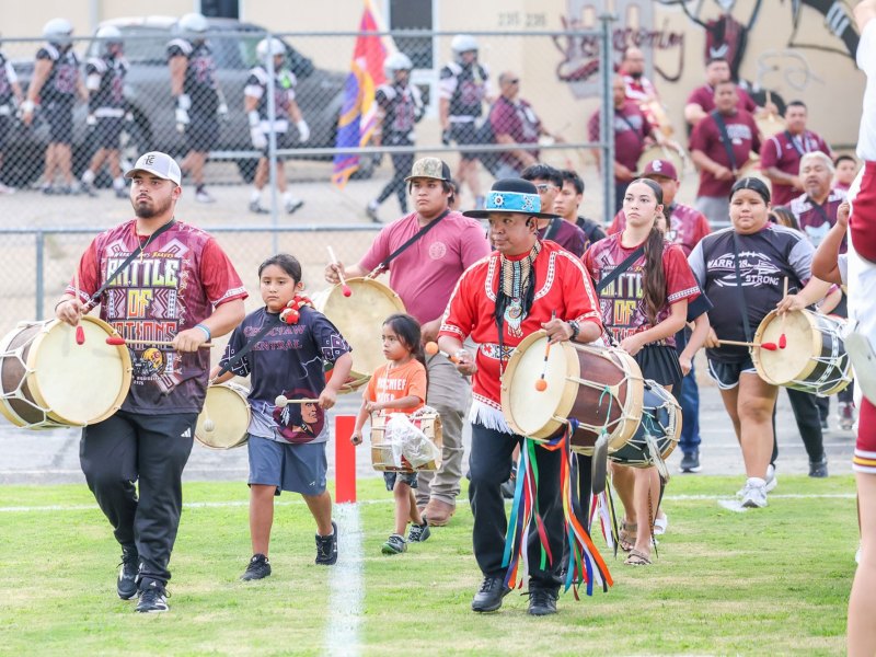 A group of people wearing red Battle of Nations shirts play drums as they walk onto a football field ahead of the football team