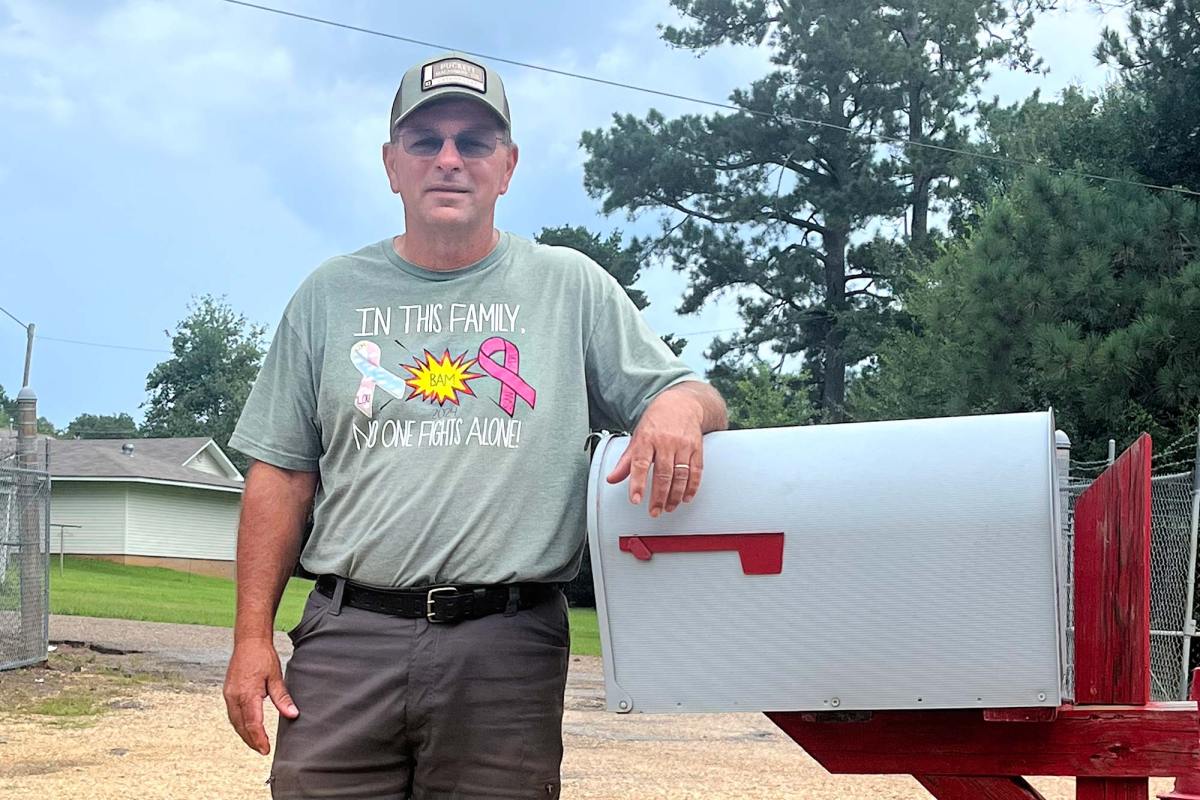 A man in a baseball cap and a shirt that reads 'In this family, no one fights alone!' poses outside against a large white and red mailbox