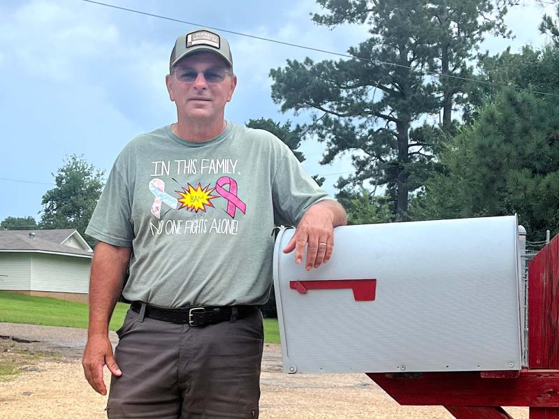 A man in a baseball cap and a shirt that reads 'In this family, no one fights alone!' poses outside against a large white and red mailbox