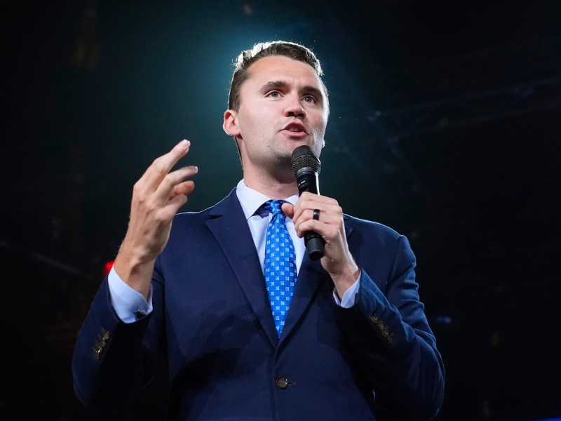 Charlie Kirk holding a microphone on a black stage