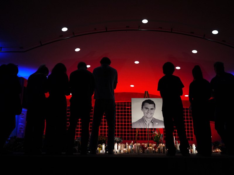 People stand around a photo of Charlie Kirk in a dark red lit room