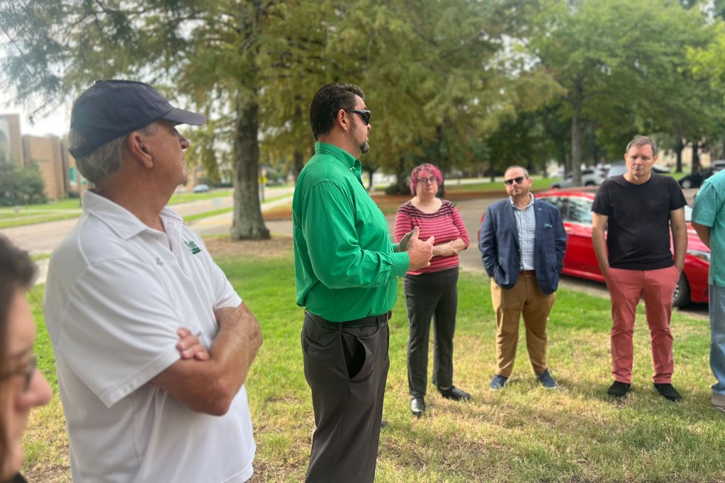 People stand outside in a loose circle as a man in a green shirt talks