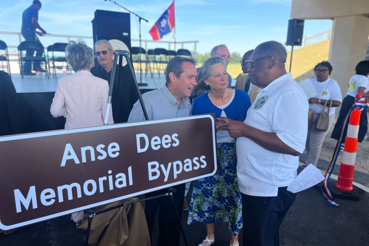 People talk at a press conference held under a bridge, a sign before them reads 'Anse Dees Memorial Bypass'